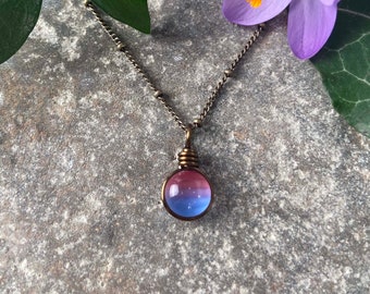 May include: A necklace featuring a small, round, translucent glass bead with a gradient of pink and blue colors. The bead is secured in a dark metal wire wrap setting and hangs from a matching dark metal chain with small, evenly spaced beads.