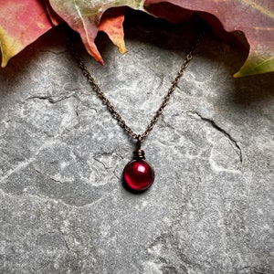 May include: A necklace featuring a small, circular, deep red glass pendant encased in dark, oxidized copper wire wrapping. The pendant hangs from a matching dark copper metal chain, creating a simple and distinct piece of jewelry.