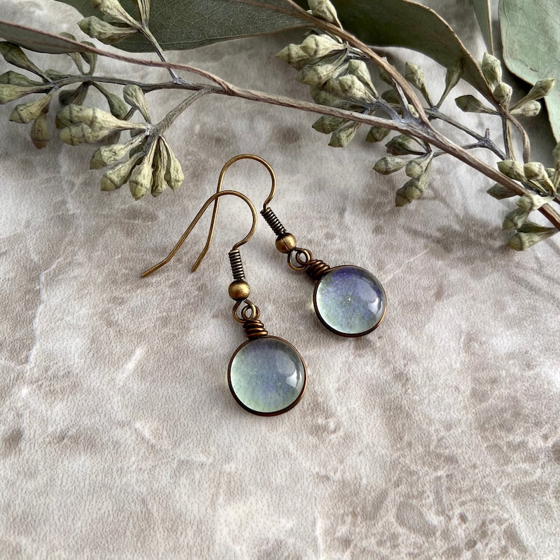 Forward facing view of the spring raindrop earrings. The raindrop is a circular dainty light green glass finished with an antique brass wire and ear wires. The angle showcases the delicate sparkle that ranges from green, blue, and purple.