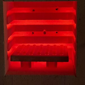 May include: The interior of a ceramic kiln glowing with intense red heat. The chamber features horizontal shelves and a central rectangular kiln shelf with a textured surface, supported by small pillars, all illuminated by the bright orange-red light.