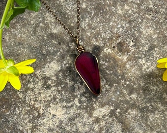 May include: A teardrop-shaped deep red pendant wrapped in dark bronze-toned wire, suspended from a matching metal chain. The pendant features a smooth, polished surface and is secured at the top with a coiled wire bail, creating a simple, elegant piece.