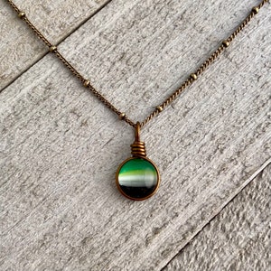 Può includere: Una collana con un ciondolo in vetro a strisce verde, bianco e nero su una catena in ottone.