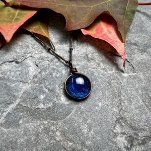 May include: A round pendant necklace featuring a dark blue, textured, glass-like center set in a bronze-toned metal frame. The pendant hangs from a matching bronze-toned chain with small, evenly spaced beads along the length of the metal links.