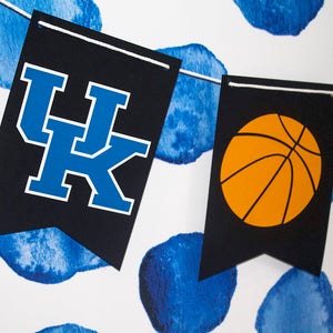 May include: A black and white banner with the letters "UK" in blue and a basketball in orange. The banner is hanging against a white background with blue watercolor circles.