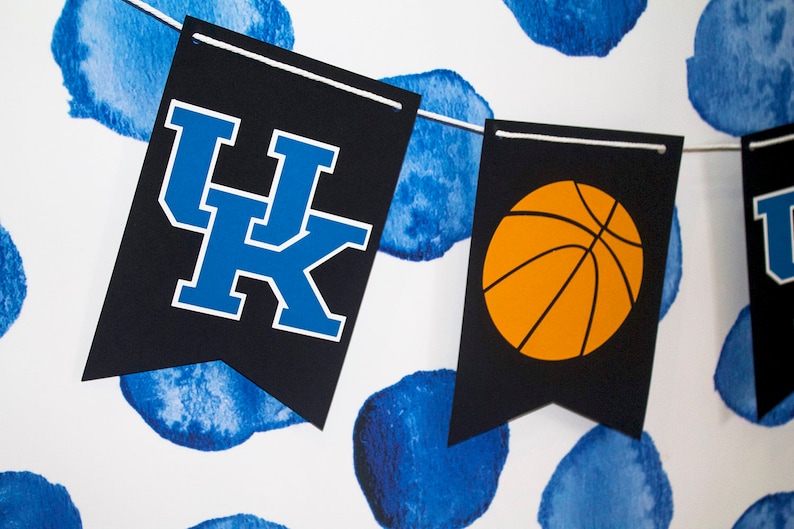 May include: A black and white banner with the letters "UK" in blue and a basketball in orange. The banner is hanging against a white background with blue watercolor circles.