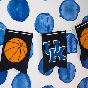 May include: A black and blue banner with the letters "UK" and a basketball graphic. The banner is strung with white string and has blue watercolor circles in the background.