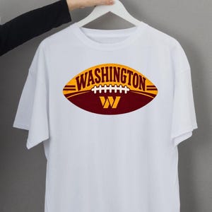 May include: A white short-sleeved t-shirt featuring a graphic of a football shape in yellow and burgundy. The word WASHINGTON is printed in dark letters across the top, with a white football lace design and a large yellow letter W below it.