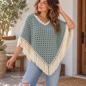 May include: A sage green crochet poncho featuring a V-neckline with a cream-colored scalloped trim and long cream fringe along the bottom edges. The garment has an open-weave diamond pattern and is paired with light blue distressed denim jeans.
