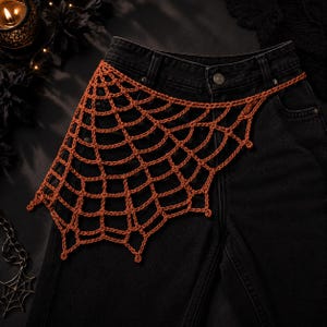 May include: A pair of black denim jeans featuring a burnt orange crocheted spider web overlay attached to the front. The web pattern is constructed from yarn and covers the upper thigh area, secured at the waistband with a single metal button.