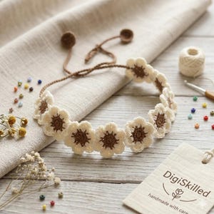May include: A crochet flower headband featuring a series of cream-colored petals with brown centers, connected by a thin brown cord with small circular ends. The accessory is displayed alongside scattered colorful beads and a wooden tag.