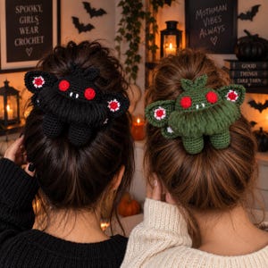 May include: Two crochet hair clips shaped like moth creatures, one in black and one in olive green. Each features large red circular eyes, small white fangs, and textured wings with white and red accents, secured in brown hair buns.