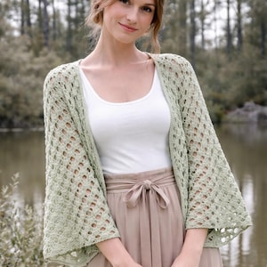 May include: A light green, open-weave crochet cardigan with wide sleeves. The cardigan is worn over a white tank top and a tan skirt with a tied waist. The background features a lake and trees.