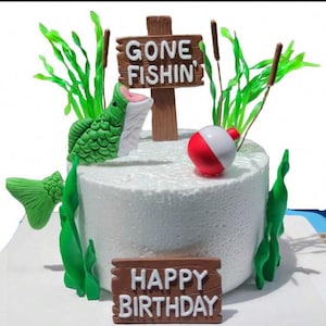 May include: A round white cake decorated with a fishing theme. It features a green fish, a red and white bobber, and a sign that says "Gone Fishin". The cake also has green seaweed and a "Happy Birthday" sign.