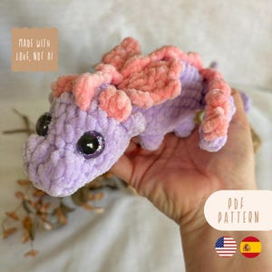 Pode incluir: Um dragão feito à mão, de crochê, em tons de lavanda e pêssego. O dragão tem olhos grandes e brilhantes e uma aparência texturizada e macia. A imagem inclui o texto "MADE WITH LOVE, NOT AI" e "PDF PATTERN".