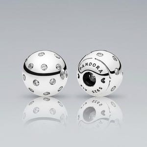 Puede incluir: Dos abalorios Pandora plateados. Cada abalorio es esférico, con una superficie pulida y acentos de cristal transparente dispersos. Un abalorio muestra el logotipo y las marcas de Pandora.