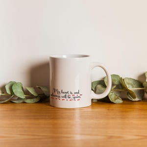 May include: A white ceramic mug with a black script that reads "My heart is, and always will be, yours." The text is accompanied by small red heart accents. The mug sits on a wooden surface with green foliage.