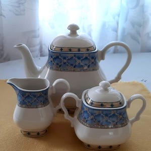 May include: A white porcelain tea set with blue and gold geometric designs. The set includes a teapot, a creamer, and a lidded sugar bowl. The teapot has a curved spout and handle. The creamer and sugar bowl have matching handles.