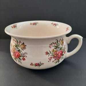 May include: A white ceramic chamber pot with a handle and floral pattern. The pot has a wide rim and is decorated with clusters of red, pink, yellow, and purple flowers and green leaves. The background is a dark grey colour.
