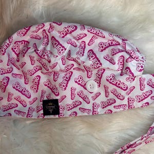 Puede incluir: Un gorro quirúrgico blanco y rosa con un patrón repetido de la palabra "Barbie". El gorro tiene un cierre de botón y lazos. Se ve una pequeña etiqueta negra con texto dorado.