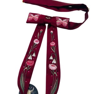 May include: A burgundy bow tie featuring embroidered pink butterflies and floral patterns. The bow tie has long, flowing tails adorned with embroidered flowers and leaves. A gold-coloured clasp secures the neckband. A small, circular tag with a skull and arrows is attached.