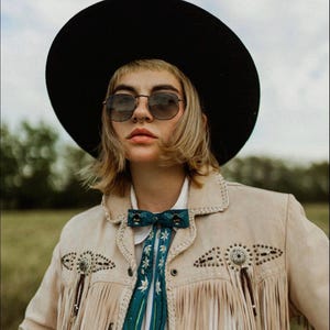 May include: A person wearing a black hat and sunglasses, a tan suede jacket with fringe and decorative metal studs, and a teal bow tie with white floral accents. The background is a field with a cloudy sky.