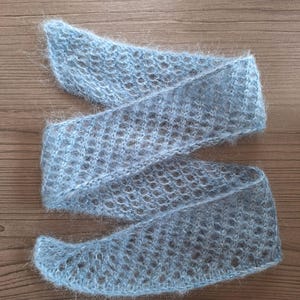 May include: A pale blue, open-knit scarf with a soft, fluffy texture. The scarf is made of a delicate, airy material and is shown against a wooden background. The scarf is approximately 178 cm long.