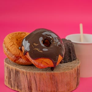 May include: A close-up of a chocolate-covered donut with a glazed donut and a small chocolate cake on a wooden stand. A paper cup with a wooden stir stick is next to the pastries against a bright pink background.