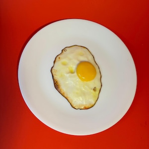 May include: A fried egg with a bright yellow yolk and a white, slightly browned egg white, sits on a white plate. The plate is centered on a vibrant red background, creating a simple, yet striking food image.
