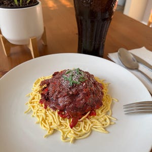 Puede incluir: Un plato de espaguetis con una generosa porción de salsa roja, adornado con hierbas y queso rallado. Un vaso alto de refresco oscuro con hielo está al lado del plato. Una pequeña planta en maceta está en el fondo.