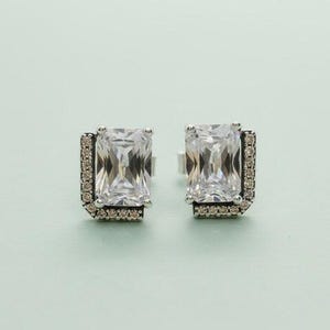 May include: A pair of silver-toned stud earrings. Each earring features a large, rectangular, clear gemstone at the center, surrounded by smaller, clear accent stones. The earrings are set against a light green background.