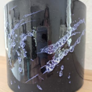 Könnte beinhalten: Ein zylindrischer Keramikbecher mit glänzend schwarzer Oberfläche und einem dekorativen Tropfenmuster in Helllila und Weiß. Die Oberfläche ist glatt und reflektierend und weist einen ausgeprägten Glasureffekt auf, der diagonal verläuft.