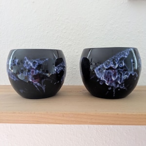 Op de afbeelding: Twee ronde keramische kommen met een glanzende zwarte afwerking, voorzien van abstracte, wolkachtige spatten van witte en paarse glazuur op de buitenoppervlakken. Deze vaten hebben een gladde, gebogen vorm en een brede opening.
