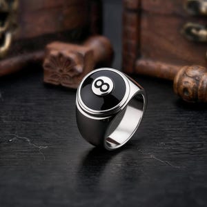 Könnte beinhalten: Ein silberfarbener Metallring mit einer kreisförmigen schwarzen Einlage auf der Vorderseite. Die Einlage zeigt die weiße Zahl Acht, die wie eine klassische Billardkugel gestaltet ist. Der Ring hat eine polierte, glatte Oberfläche.