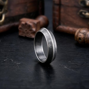 Può includere: Un anello color argento con una finitura scura, usurata e testurizzata. La fascia presenta un bordo centrale liscio e rialzato che corre orizzontalmente lungo la circonferenza, creando un sottile contrasto con la superficie opaca e scura.