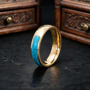 May include: A polished gold-tone ring featuring a smooth, rectangular inlay of bright turquoise stone with dark, natural veining. The band has a rounded profile and a high-shine metallic finish, showcasing a clean, simple design with a vibrant blue accent.