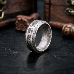 May include: A silver-toned metal ring featuring an oxidized finish with engraved details. The band displays a repeating Celtic knot pattern, a tree motif, and a series of runic characters encircling the outer surface of the circular band.