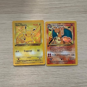 Puede incluir: Dos cartas coleccionables de Pokémon. Una presenta "Ditto Pikachu" con una ilustración de Pikachu amarillo. La otra muestra "Ditto Charizard" con una ilustración de Charizard naranja. Ambas cartas tienen texto e ilustraciones.