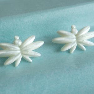 Könnte beinhalten: Ein Paar weiße Ohrstecker mit einem mehrblättrigen Blumendesign und Perlmuttglanz. Jeder Ohrring besteht aus mehreren länglichen, spitz zulaufenden Blütenblättern, die von einer kleinen zentralen Kugel ausgehen und eine sternförmige Struktur bilden.