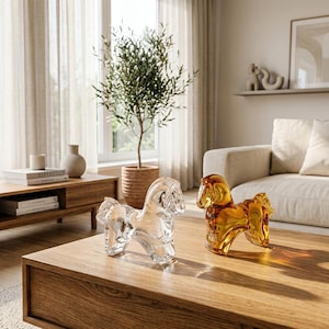 May include: Two glass horse figurines, one clear and one amber, sit on a wooden coffee table. The clear horse is in the foreground, while the amber horse is to the right. The room has a neutral color palette with a plant and a sofa in the background.