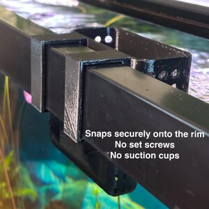 May include: Black plastic aquarium rim clip. The clip has a rectangular shape with a curved section and several small holes. Text on the image reads "Snaps securely onto the rim. No set screws. No suction cups."