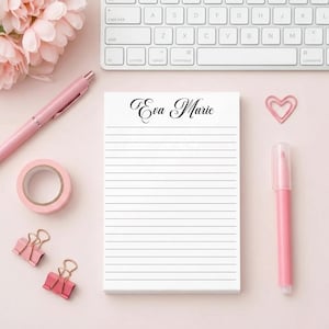 May include: A notepad with the name "Eva Marie" in elegant script, surrounded by pink office supplies. A pink pen, tape, paper clips, and a marker are arranged on a light pink surface, with a white keyboard and pink flowers in the background.