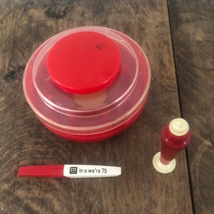 May include: A red and clear plastic container with a red lid, a red and white pen with the text "in 76 we're 75", and a red and white bottle stopper with a white base are on a wooden surface.