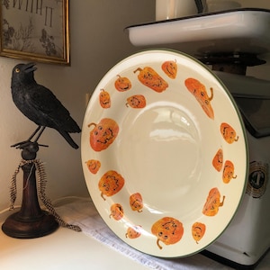 May include: A cream-colored plate with a green rim, decorated with orange jack-o'-lanterns. A black crow figurine sits on a decorative stand to the left. A vintage scale is in the background.