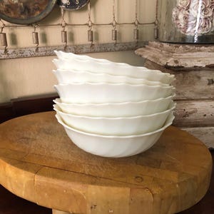 May include: A stack of six white, scalloped-edge bowls. The bowls are stacked on a round, wooden cutting board. The background includes antique clocks and decorative items.