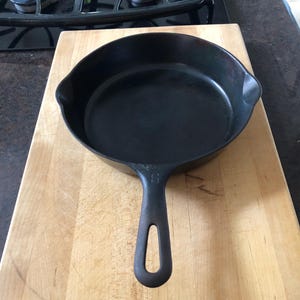 May include: A black cast iron skillet with a long handle and a rounded cooking surface. The skillet is sitting on a light-colored wooden cutting board. The number "5" is visible on the handle.