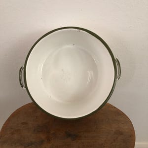 May include: A white enamel bowl with a dark green rim and handles. The bowl is sitting on a wooden stool. The bowl has a simple, classic design.