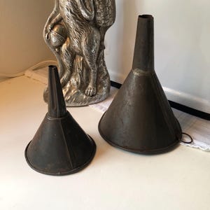 May include: Two antique metal funnels. The funnels are dark brown with a conical shape. One funnel is smaller than the other. The funnels are likely used for pouring liquids or powders into containers. The funnels are placed on a white surface.