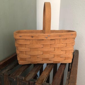 May include: A light brown, rectangular woven basket with a tall, arched handle. The basket is made of thin wooden strips, with metal rivets along the top edge. It sits on a weathered wooden rack.