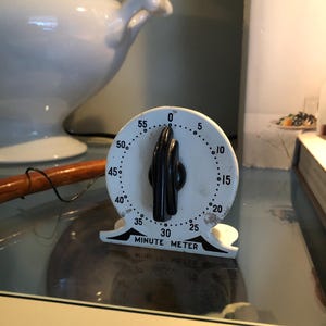 May include: A vintage white Minute Meter timer with a black handle, displaying the time. The timer has a circular face with numbers and markings. The text "MINUTE METER" is visible. A white soup tureen is in the background.