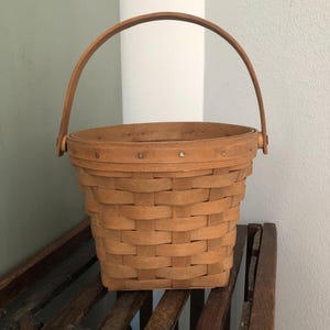May include: A light brown woven basket with a curved handle. The basket has a round opening and a rectangular base. The handle is attached to the sides of the basket. The basket is sitting on a dark brown wooden rack.
